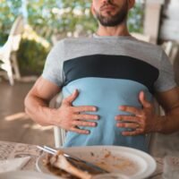A man sits back feeling full after dining, showing a satisfied expression while touching his stomach.